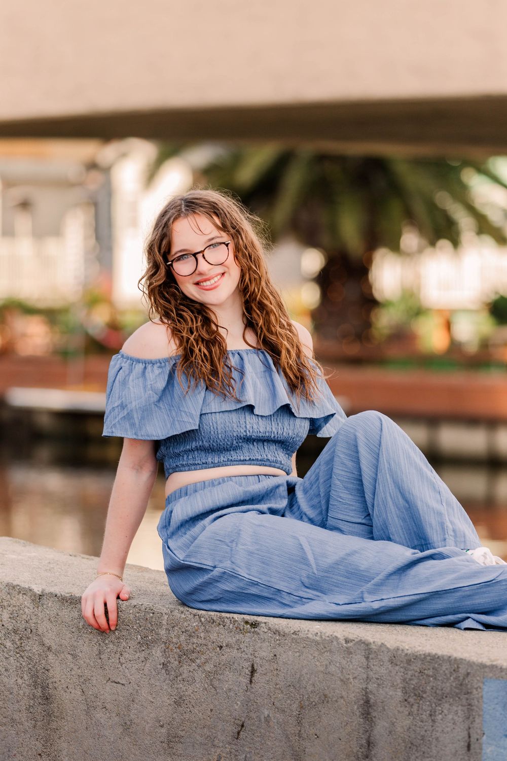 Casual outdoor portrait in a blue two-piece outfit sitting on concrete ledge near palm trees.