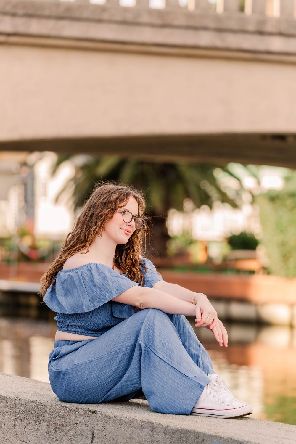 Casual summer portrait sitting on concrete ledge wearing denim two-piece set and white sneakers.