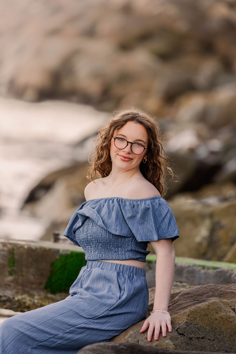 A woman in a blue chambray dress sits on coastal rocks during an outdoor photo session.