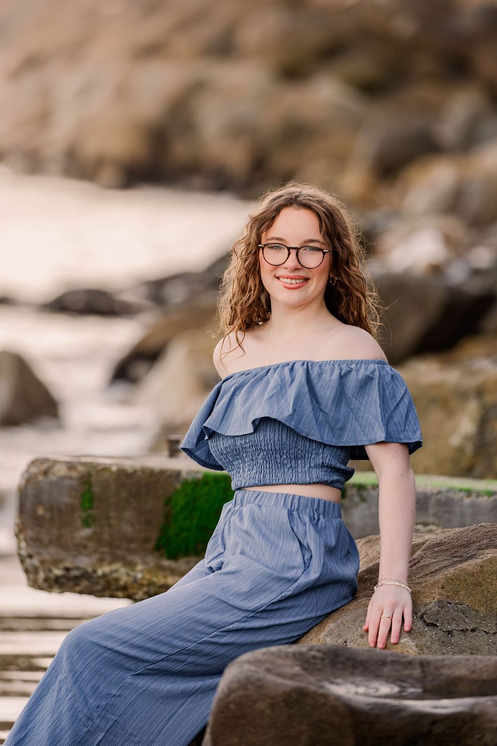 A maternity photo series shows a woman in a blue denim dress sitting on coastal rocks at the beach.