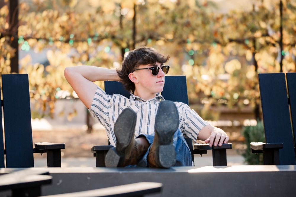 Person wearing sunglasses sits at outdoor table with laptop and coffee cup in autumn setting.