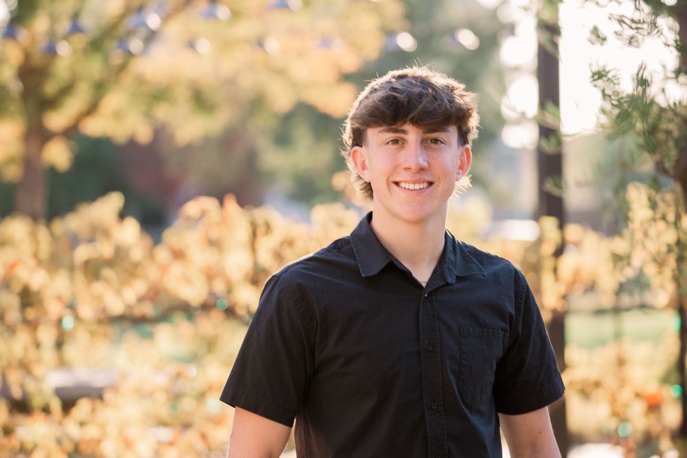 A young person in a black shirt poses outdoors during golden hour with autumn foliage in the background.