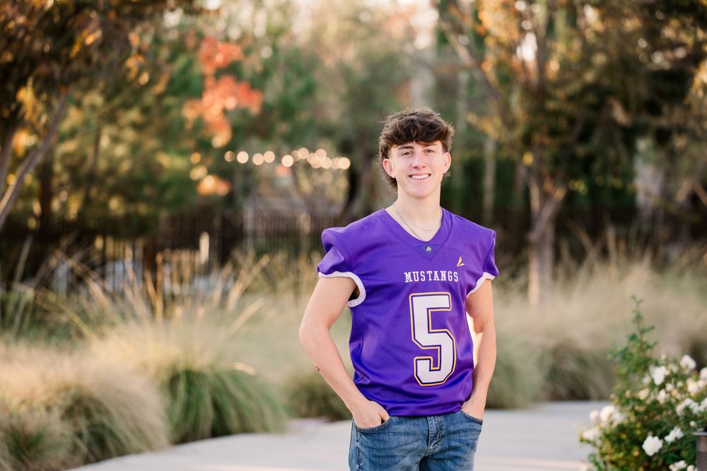 High school football player in purple jersey stands in outdoor setting with blurred natural background.