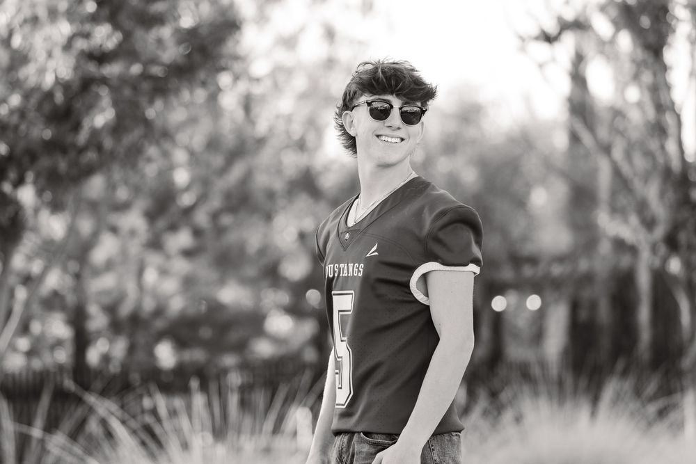 Black and white photo of a person wearing sunglasses and a v-neck t-shirt smiling outdoors.