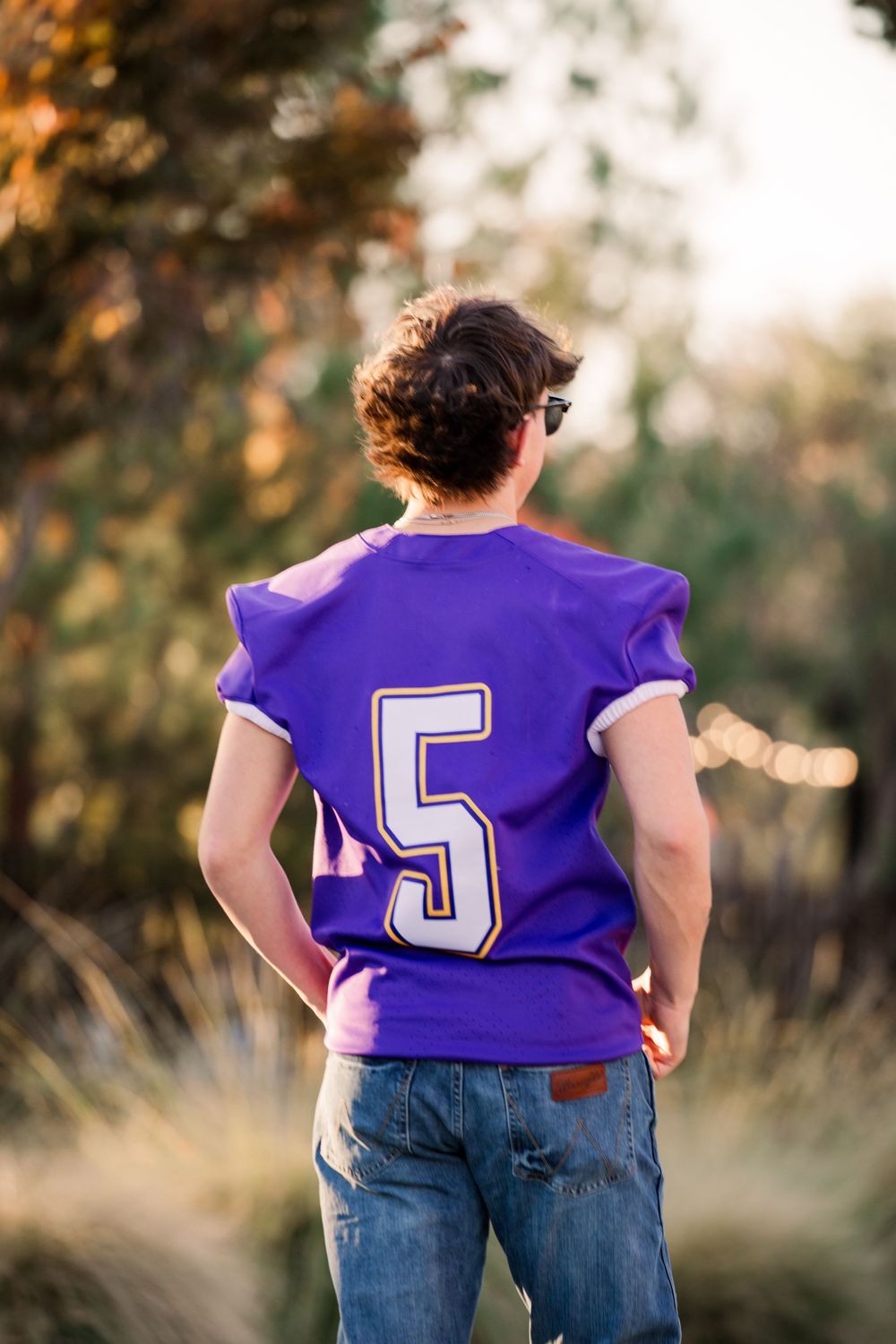 Back view of someone wearing a purple sports jersey with number 5 and blue jeans in golden sunlight.
