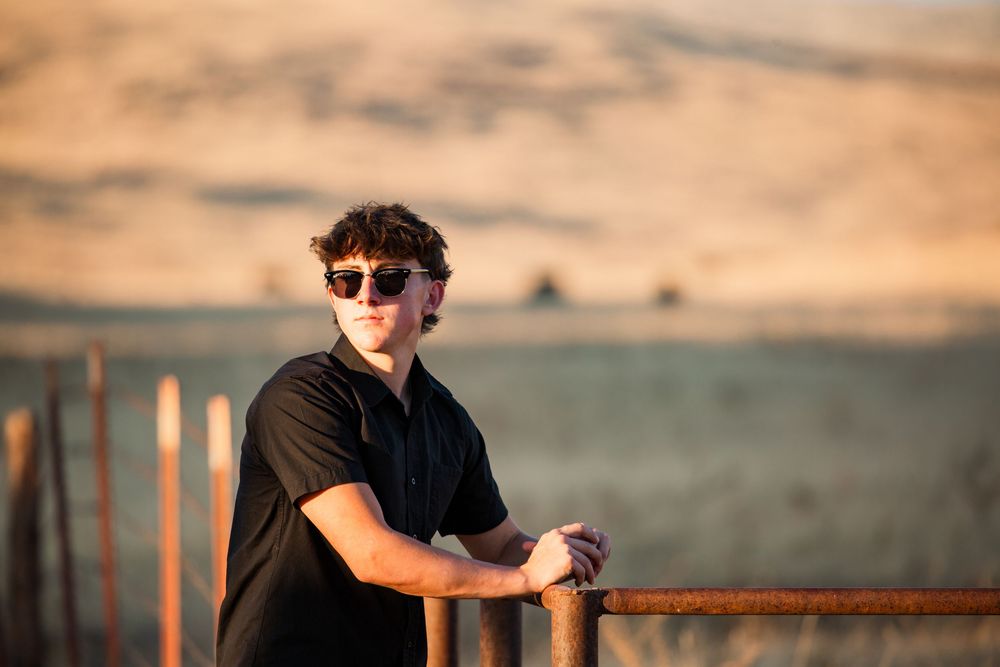 Person in sunglasses leans on wooden railing at sunset with desert landscape in background.
