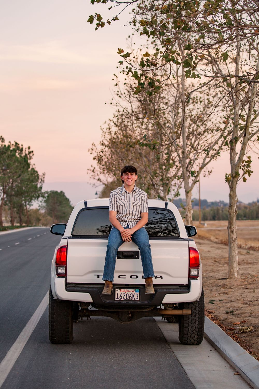 Person sitting on the tailgate of a white Toyota pickup truck at sunset with trees in the background.