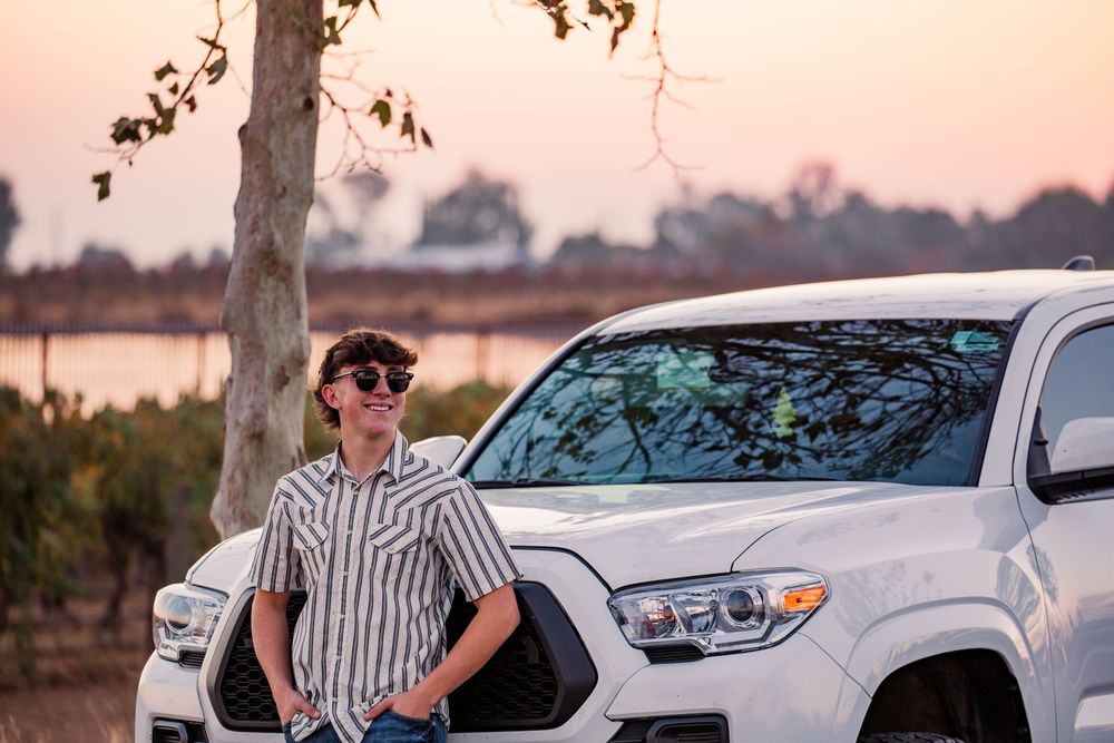 Person in striped shirt leans against white car during sunset with trees in background.