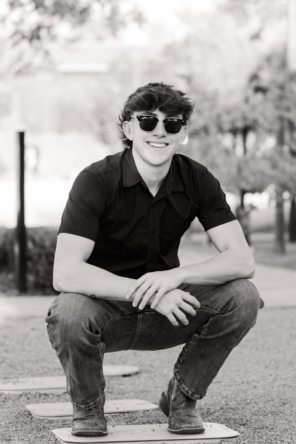 Black and white portrait of a young person in sunglasses and black polo shirt crouching outdoors.