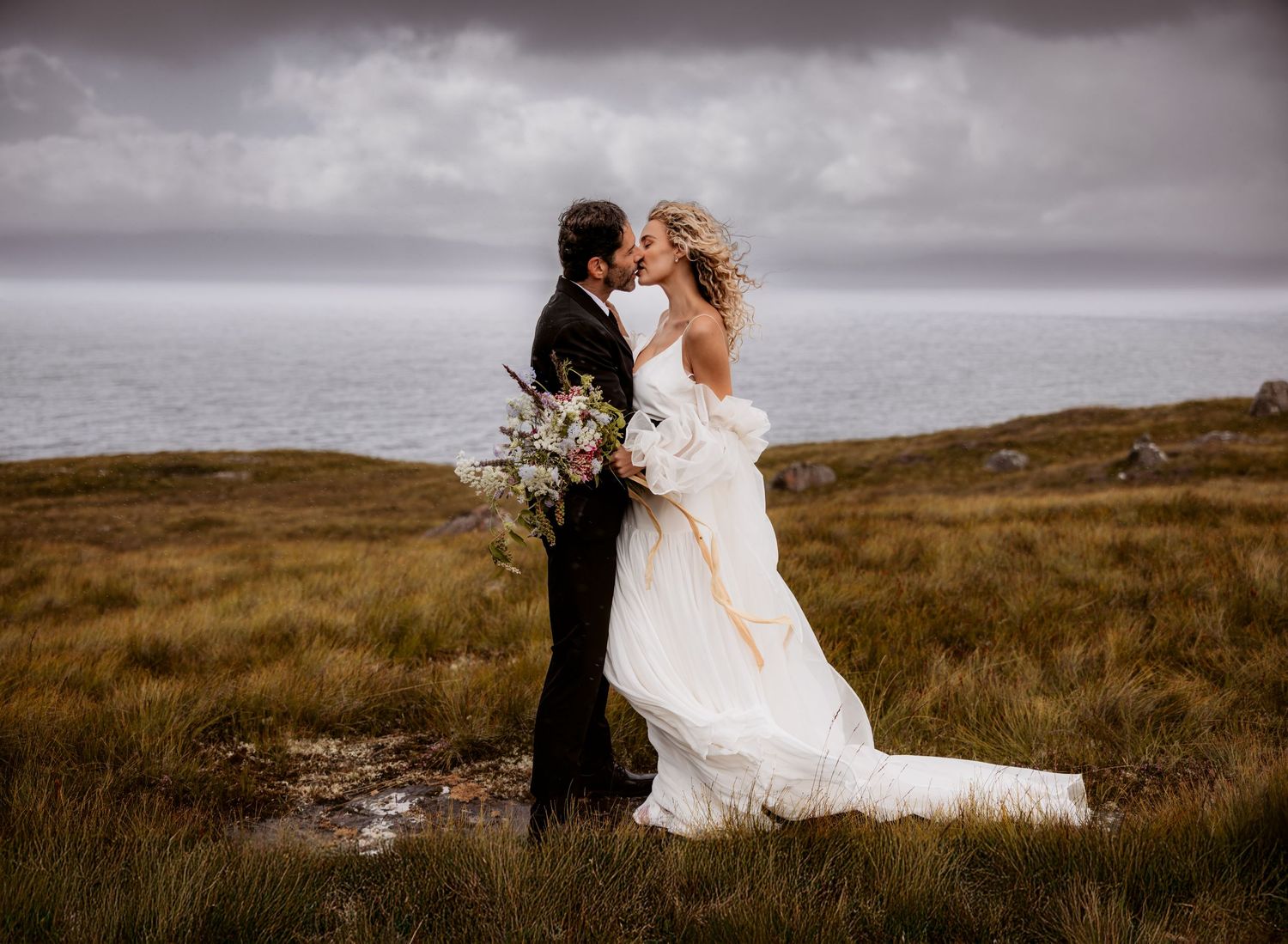 A couple embraces on a windswept hilltop during their wedding, with stormy skies and rugged landscape in the background.