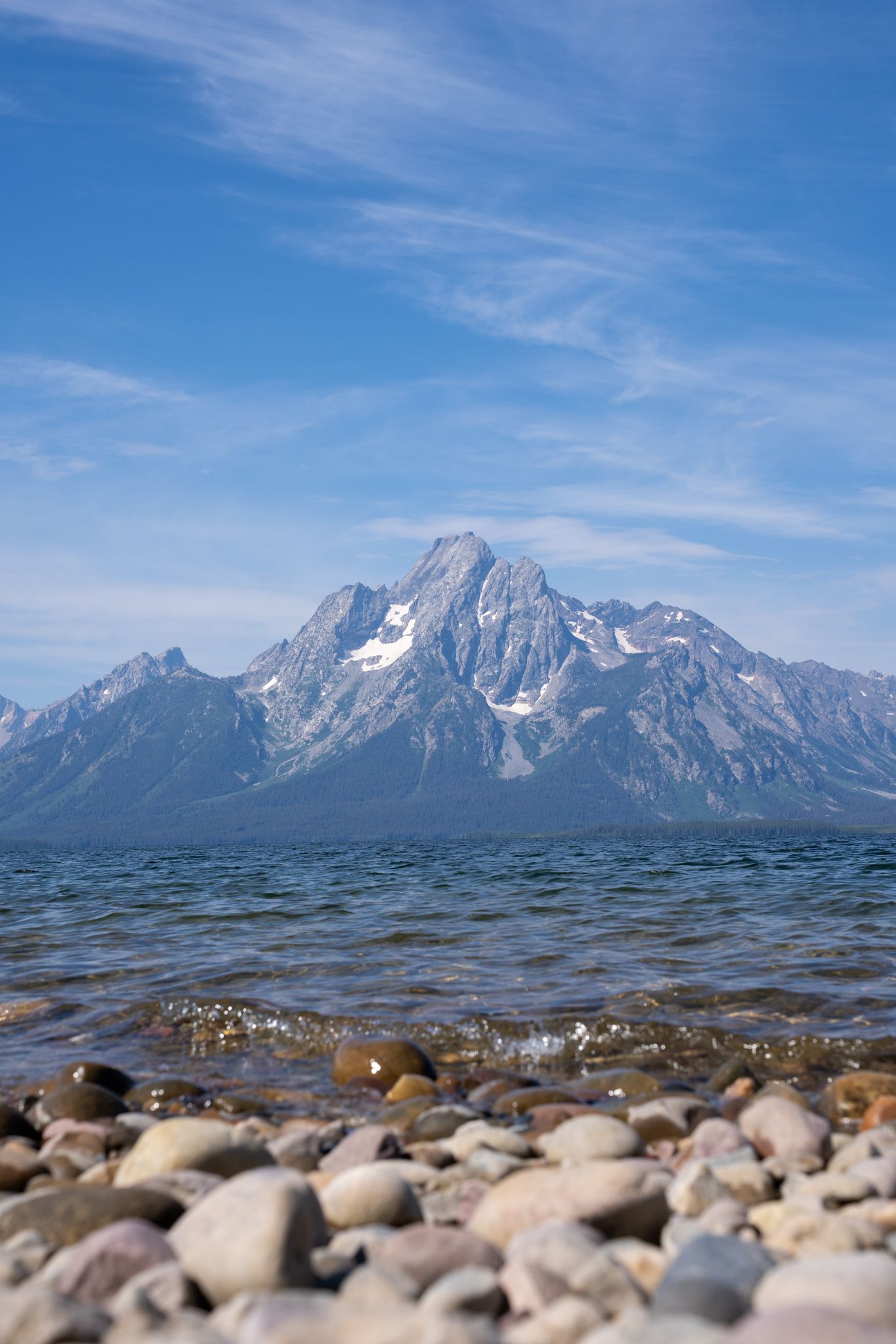 Snow-capped mountain peaks rise above Jackson Lake with smooth stones visible along the shoreline under blue skies.
