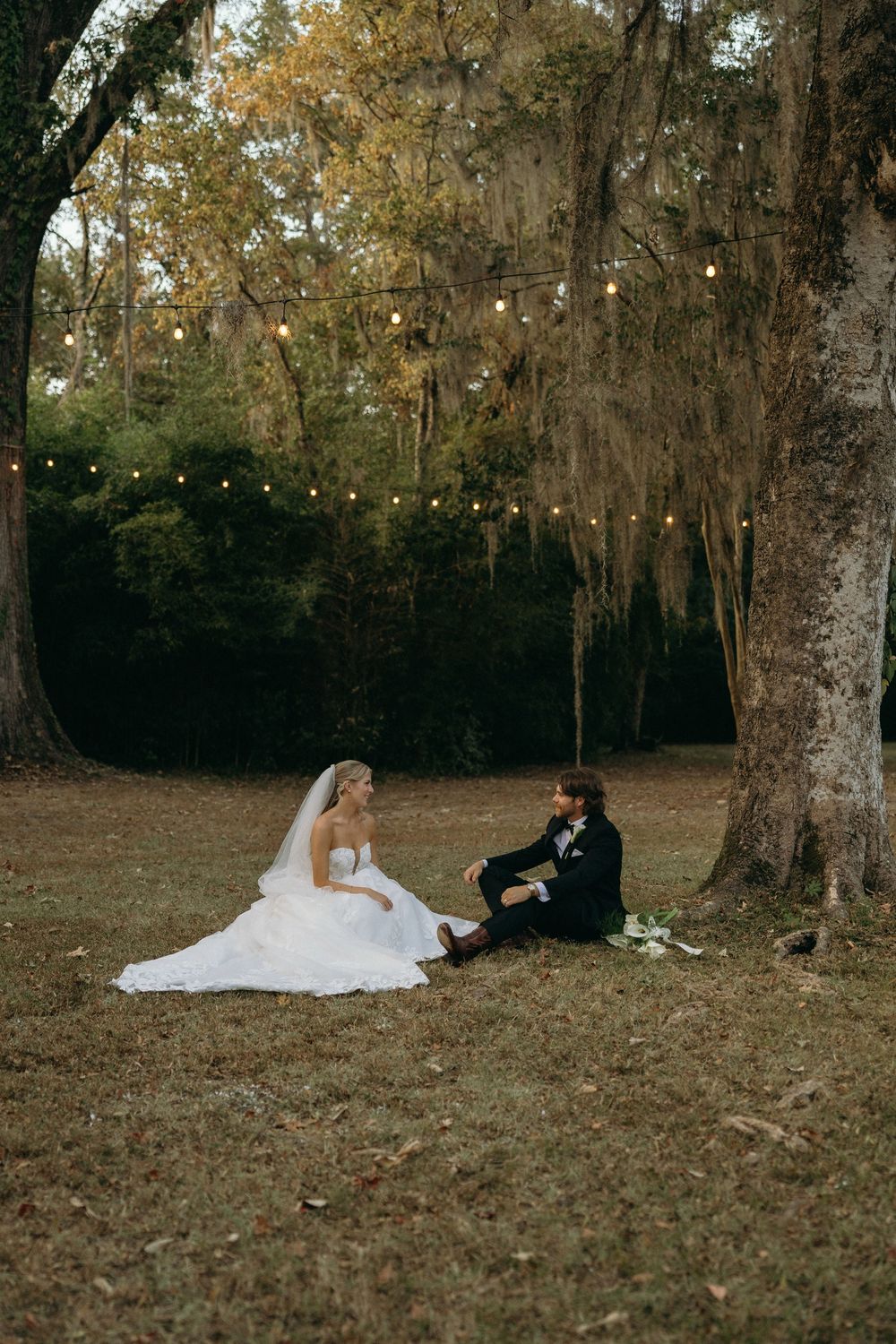 A couple in wedding attire rests together on grass beneath moss-draped oak trees in an autumn setting at The Elms of Coosada in Coosada, Ala