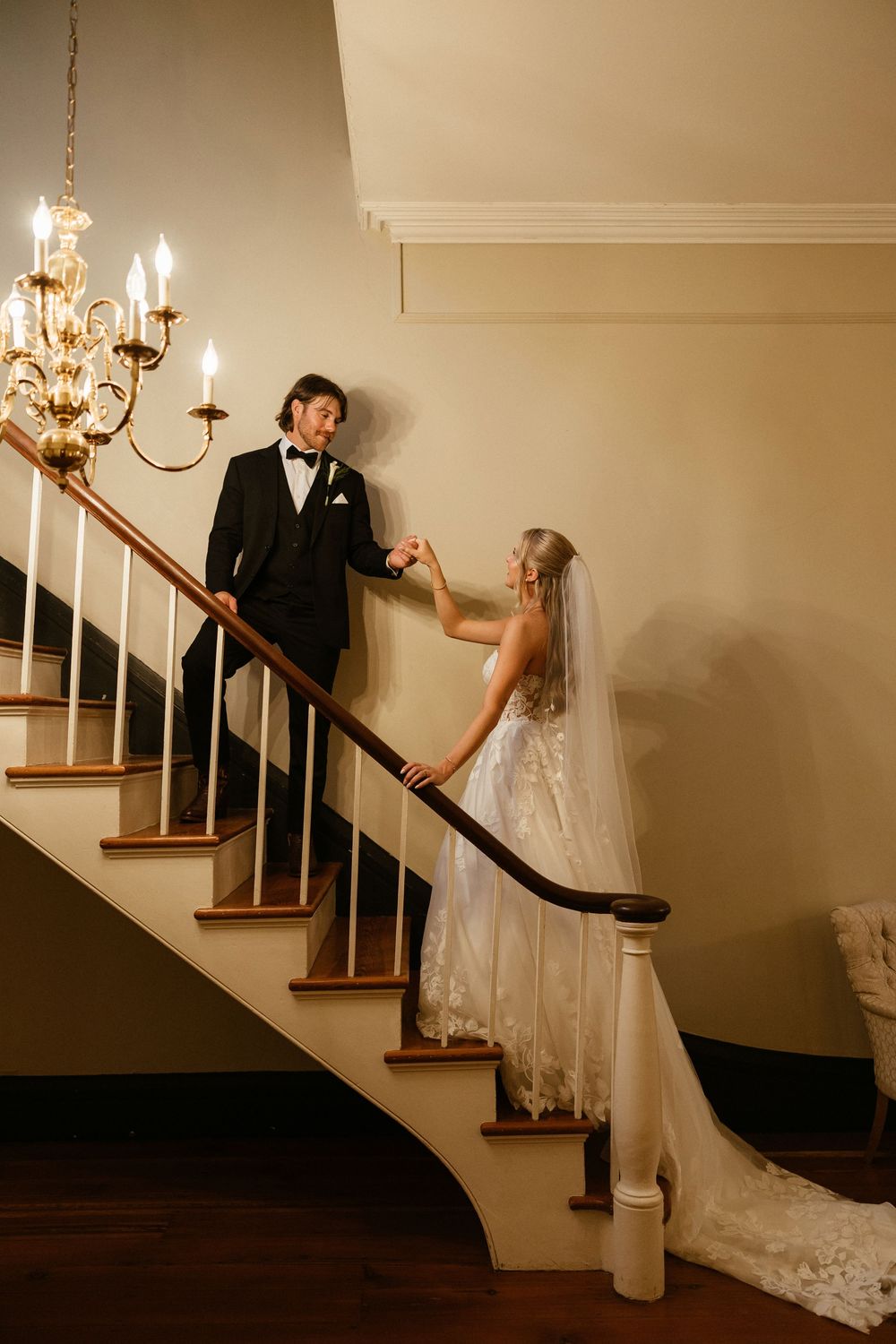 A creative wedding photo showing a couple playfully posing on a staircase with a chandelier overhead at The Elms of Coosada in Coosada