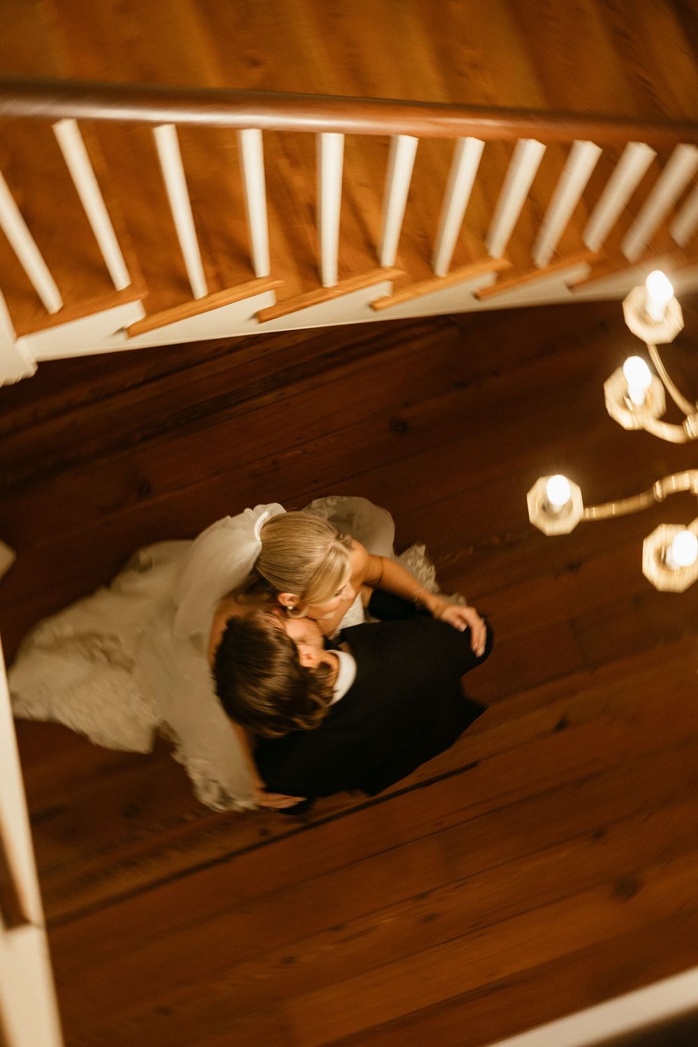 Overhead view of a romantic couple dancing on wooden floors beneath warm string lights and white staircase railings at The Elms of Coosada