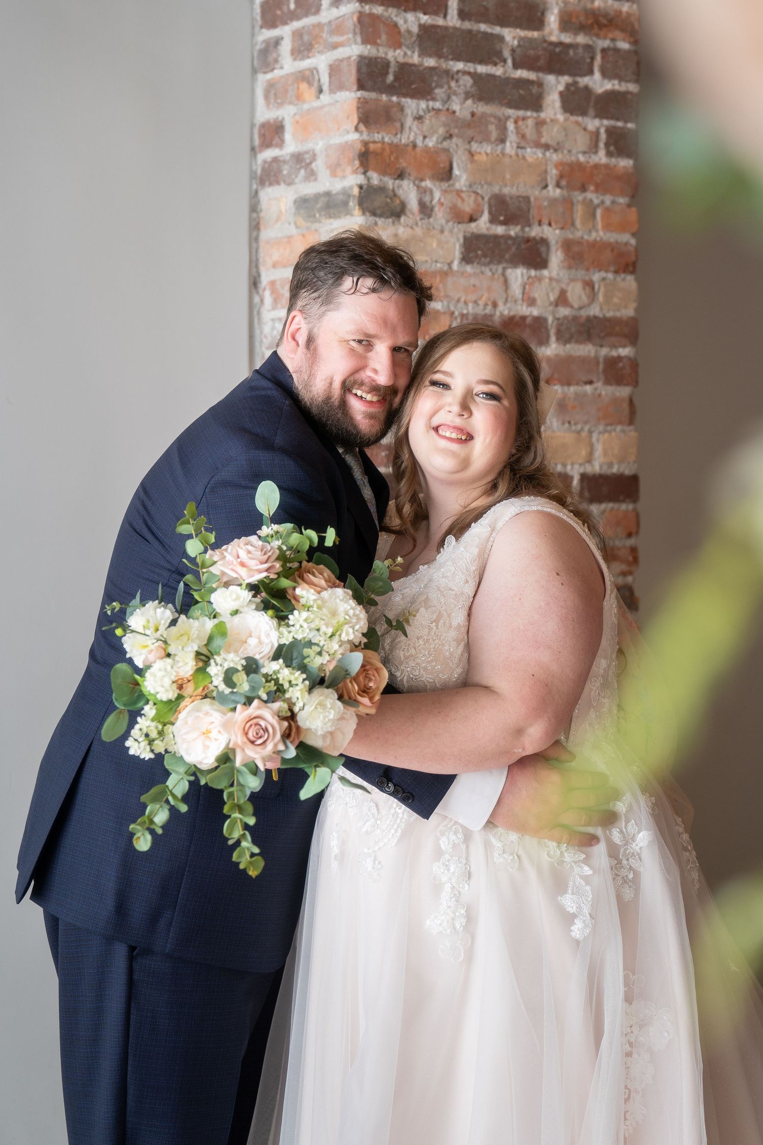 A happy couple shares a tender moment against a brick wall backdrop, with a beautiful bouquet of blush roses and greenery.