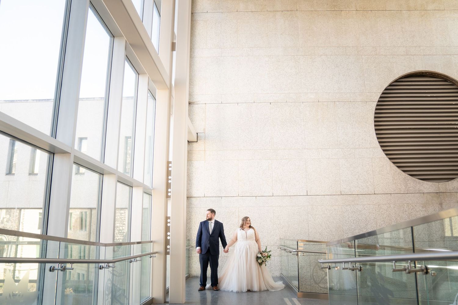 A couple walks together in a modern building with large windows and a circular vent on the wall.