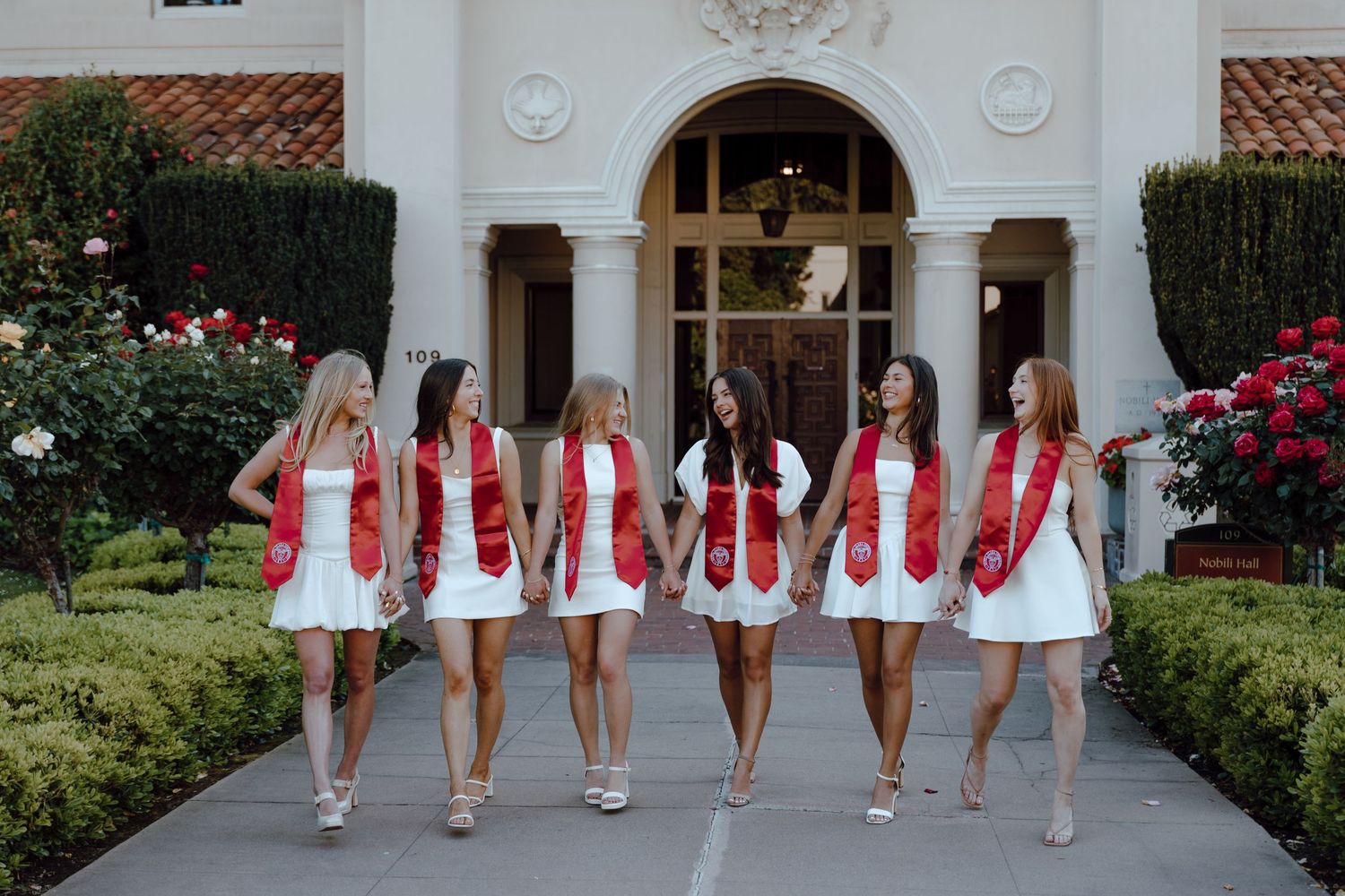 Group of Santa Clara University graduates hold hands and walk together, with rows of roses. Taken by Bay Area Graduation Photographer.