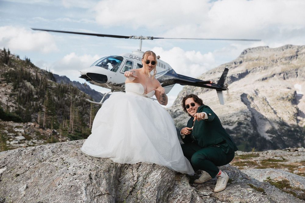 Bride and friend pose with thumbs up on rocky mountain summit with helicopter in background.