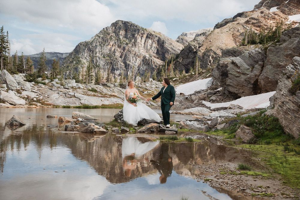 Couple shares romantic moment by reflective alpine lake with snow-dusted mountain backdrop.