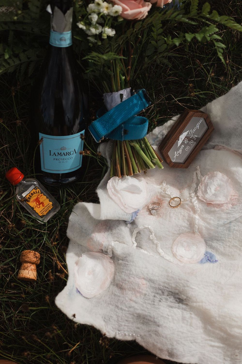 Wedding flat lay featuring LaMarca prosecco, bridal bouquet, rings, and accessories on white cloth.