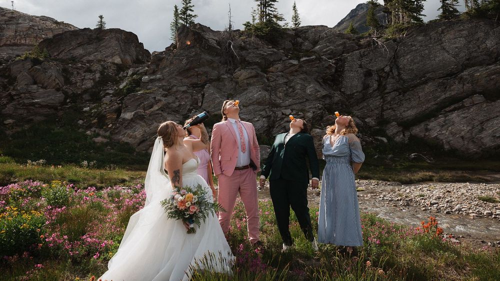 Wedding party of five stands among wildflowers with dramatic rocky mountain cliffs behind them.