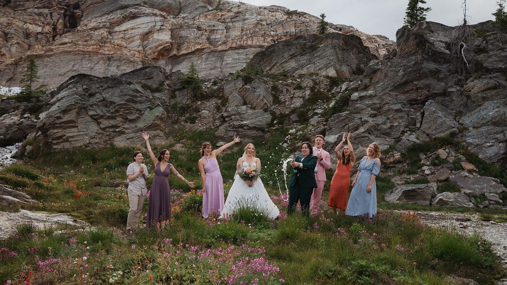Bridal party celebrates with raised arms among colorful wildflowers against rugged rocky cliffs.