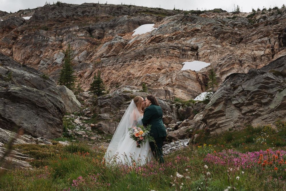 Bride and groom share a kiss among wildflowers with dramatic rocky mountain face behind them.