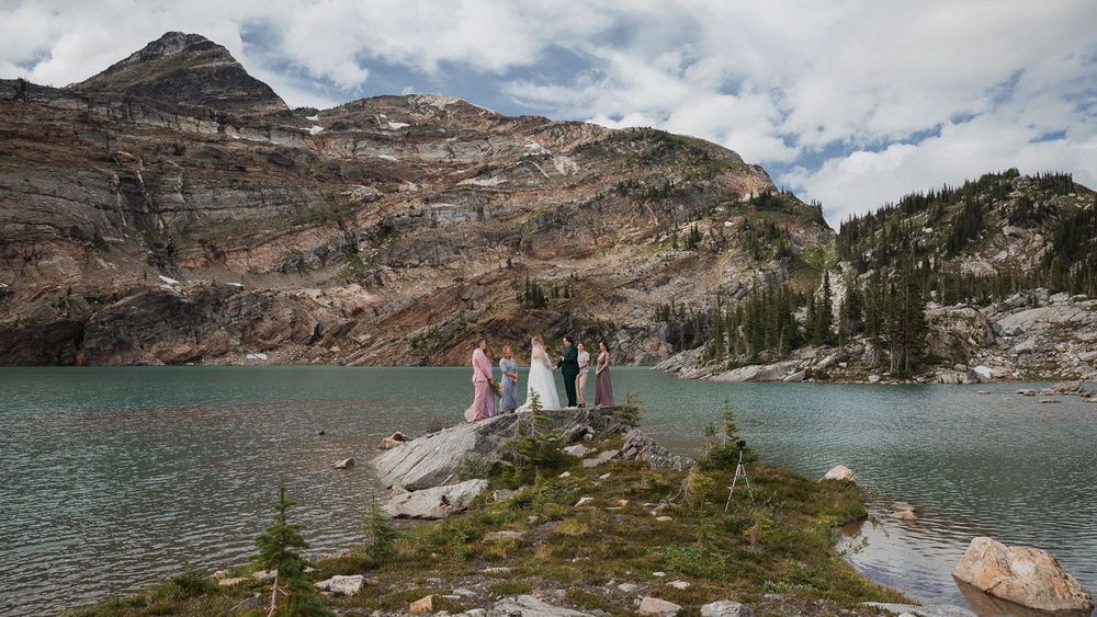 Wedding party stands on rocky shore of stunning turquoise alpine lake surrounded by mountains.