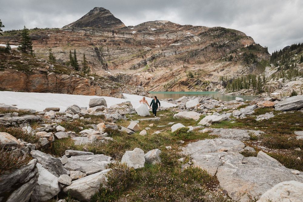 Person in teal jacket hikes across rocky alpine landscape with snow patches and turquoise lake.