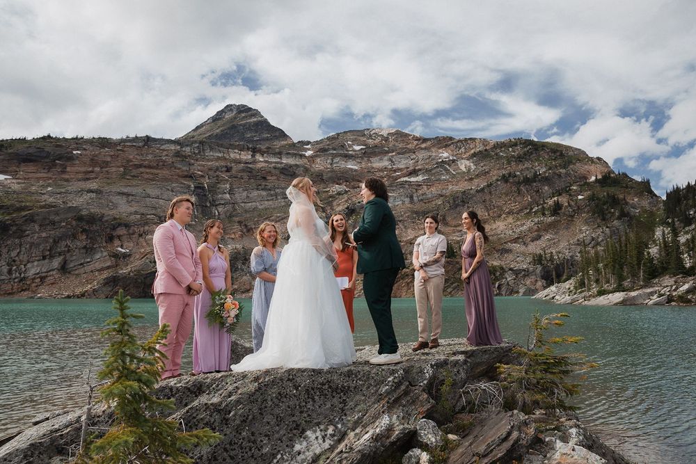 Wedding party stands by turquoise alpine lake during outdoor mountain elopement ceremony.