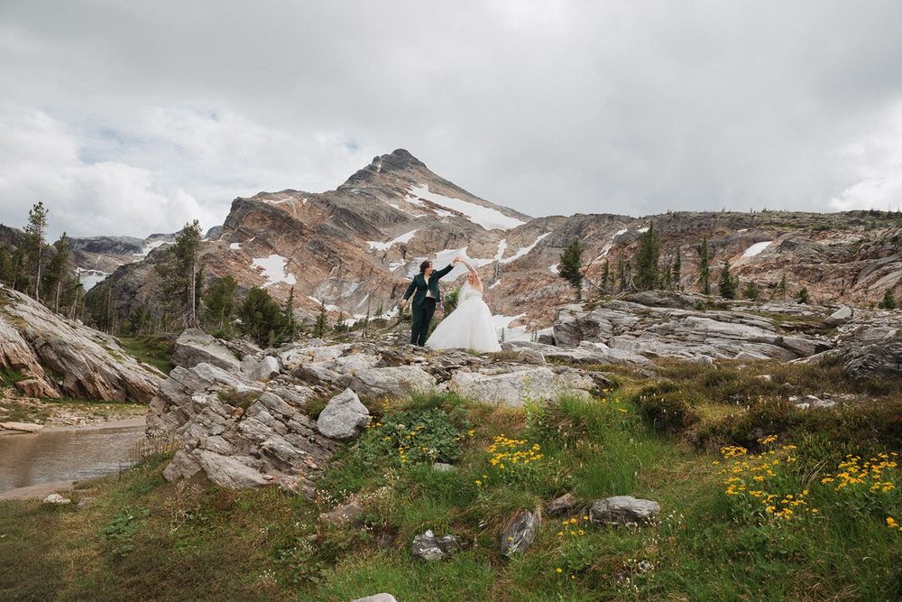 Bride and groom embrace among wildflowers with dramatic snow-covered mountain peak behind them.