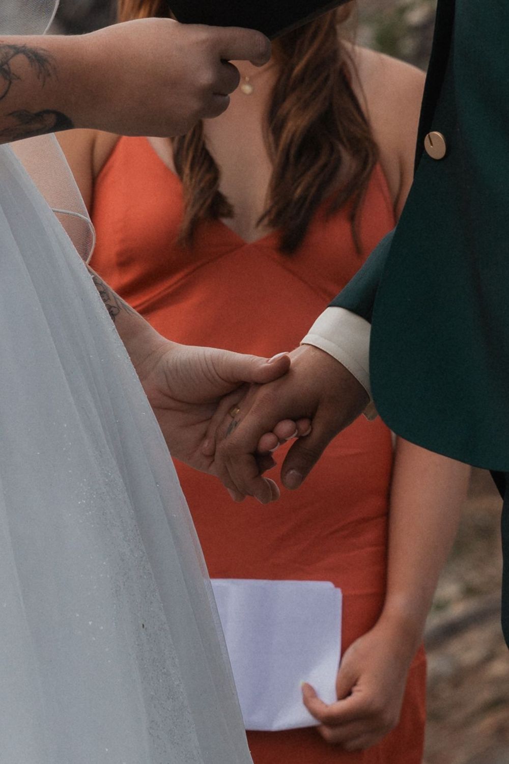 Couple holds hands during wedding ceremony while officiant in orange dress reads vows.