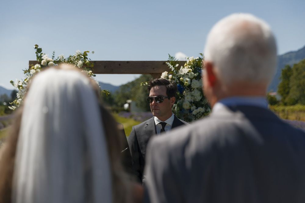 Groom in sunglasses stands at outdoor wedding arch with floral arrangements, mountains visible behind him on a sunny day.