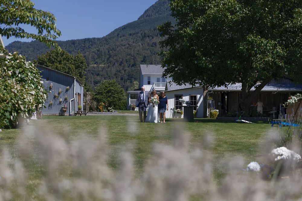 Couple walks across lush green lawn with mountain and white farmhouse buildings visible in background on a sunny day.