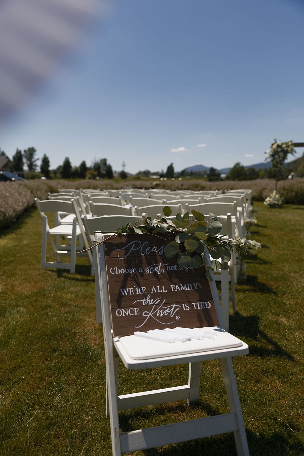 Wooden wedding sign reading we are all family once the knot is tied on white chair with rows of seats behind.