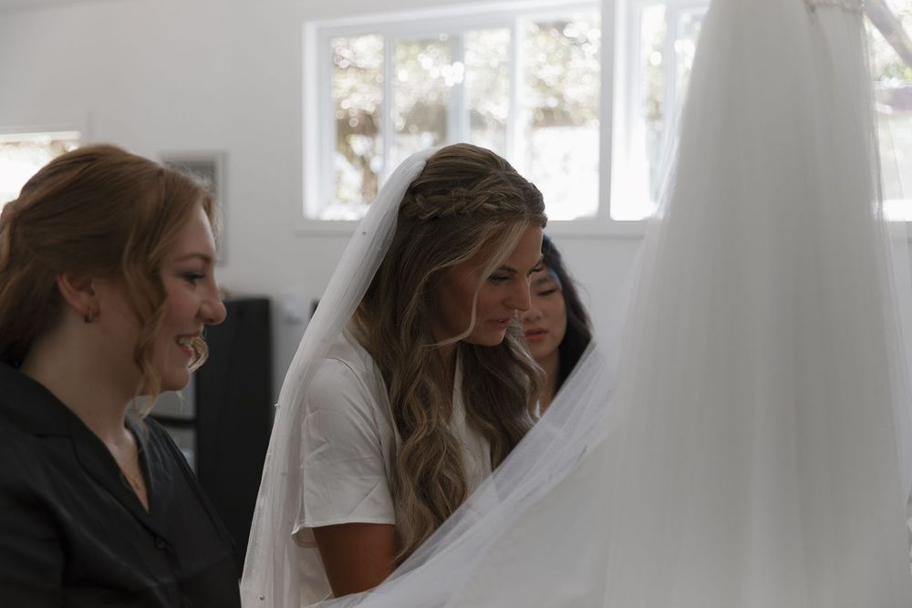 Bride and bridesmaid smiling while examining a white wedding veil in a bright dressing room.