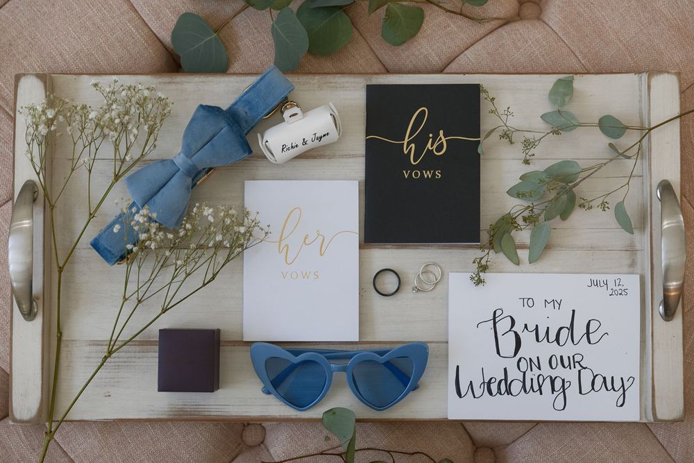 Flat lay of wedding day details including vow books, blue bow tie, heart sunglasses, and bridal card.