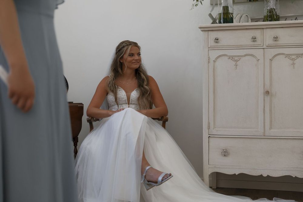 Bride in a beaded wedding gown sitting and smiling in a bright white dressing room.