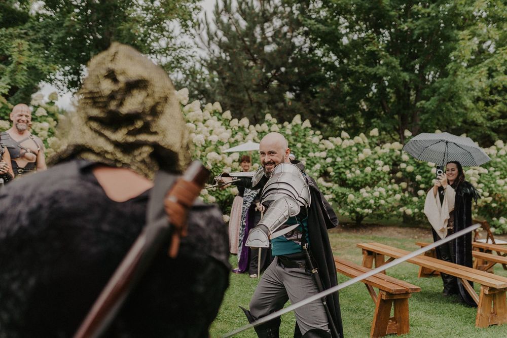 Man in medieval knight costume engaged in a sword fight at an outdoor fantasy-themed wedding celebration.