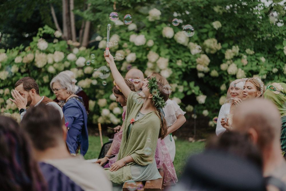 Wedding guest in green dress joyfully raising her arm among guests at an outdoor garden ceremony.