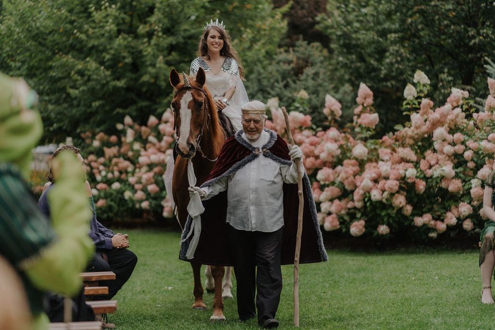 Bride wearing a crown arriving on horseback led by a knight at a medieval fantasy wedding.