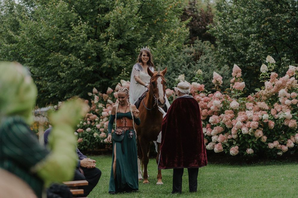 Bride on horseback in white gown with crown greeted by guests at a medieval-themed outdoor wedding.