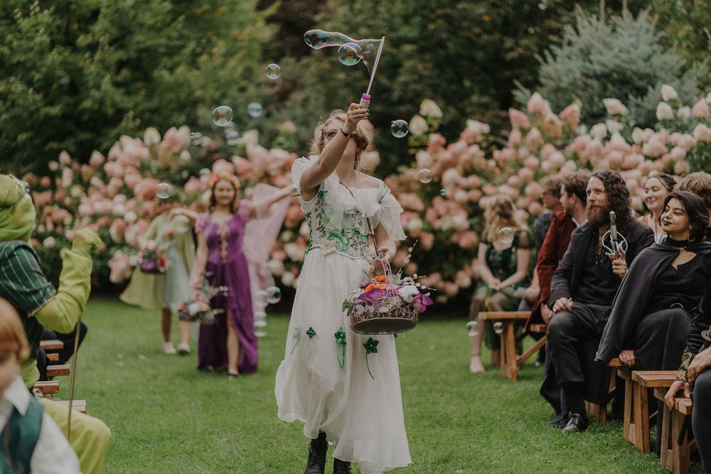 Flower girl in white dress blowing bubbles while walking down the aisle at an outdoor garden wedding.
