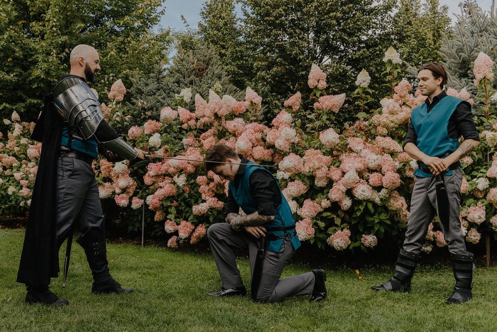 Three people in medieval fantasy costumes acting out a kneeling scene near pink hydrangeas.
