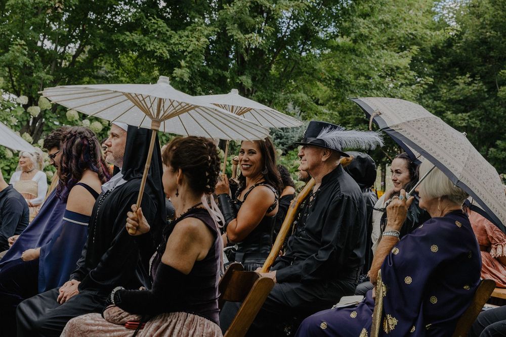 Wedding guests in medieval costumes sit with parasols at an outdoor garden ceremony.