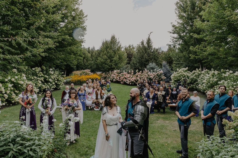 Bride in white gown and groom in knight armor stand at outdoor medieval themed wedding ceremony.