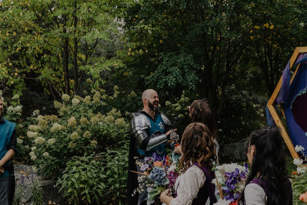Knight in armor greets guests holding colorful bouquet at outdoor medieval fantasy wedding.