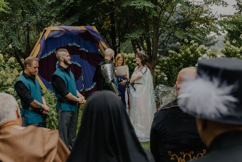 Couple exchanges vows at medieval themed outdoor wedding ceremony under a purple decorative arch.