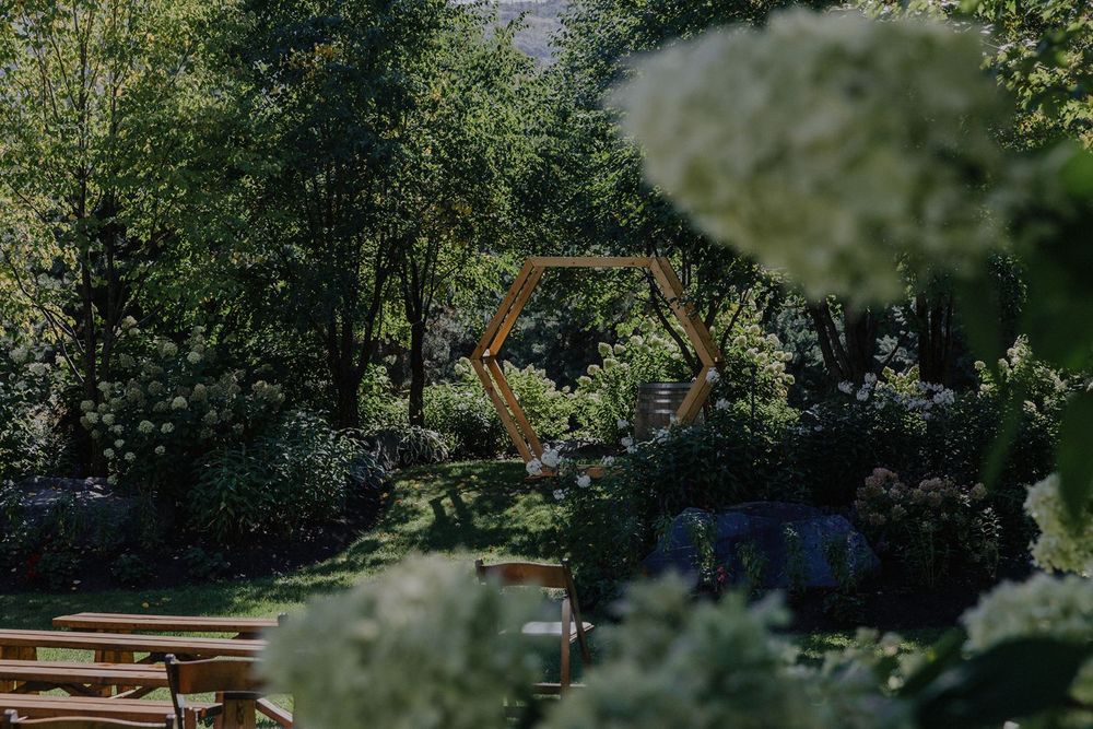 Wooden hexagonal wedding arch framed by lush green trees and white hydrangeas in garden setting.