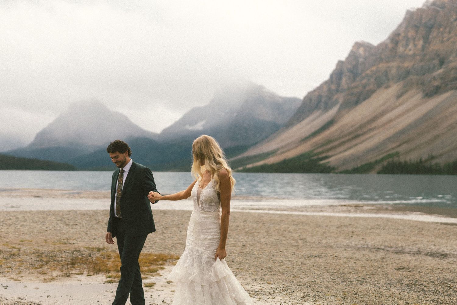 bride and groom walk hand and hand along the beach of a mountain lake in Kananaskis